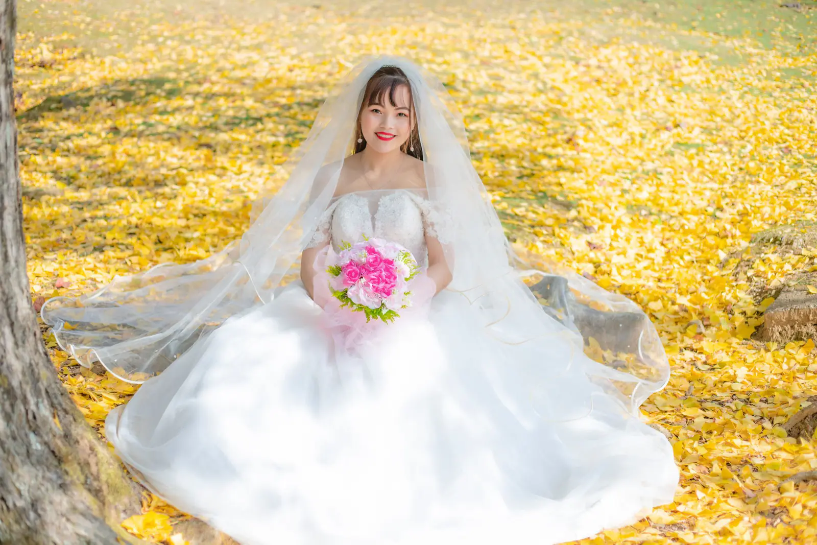 Bride in a white gown and veil sitting among golden leaves, holding a bouquet, smiling.