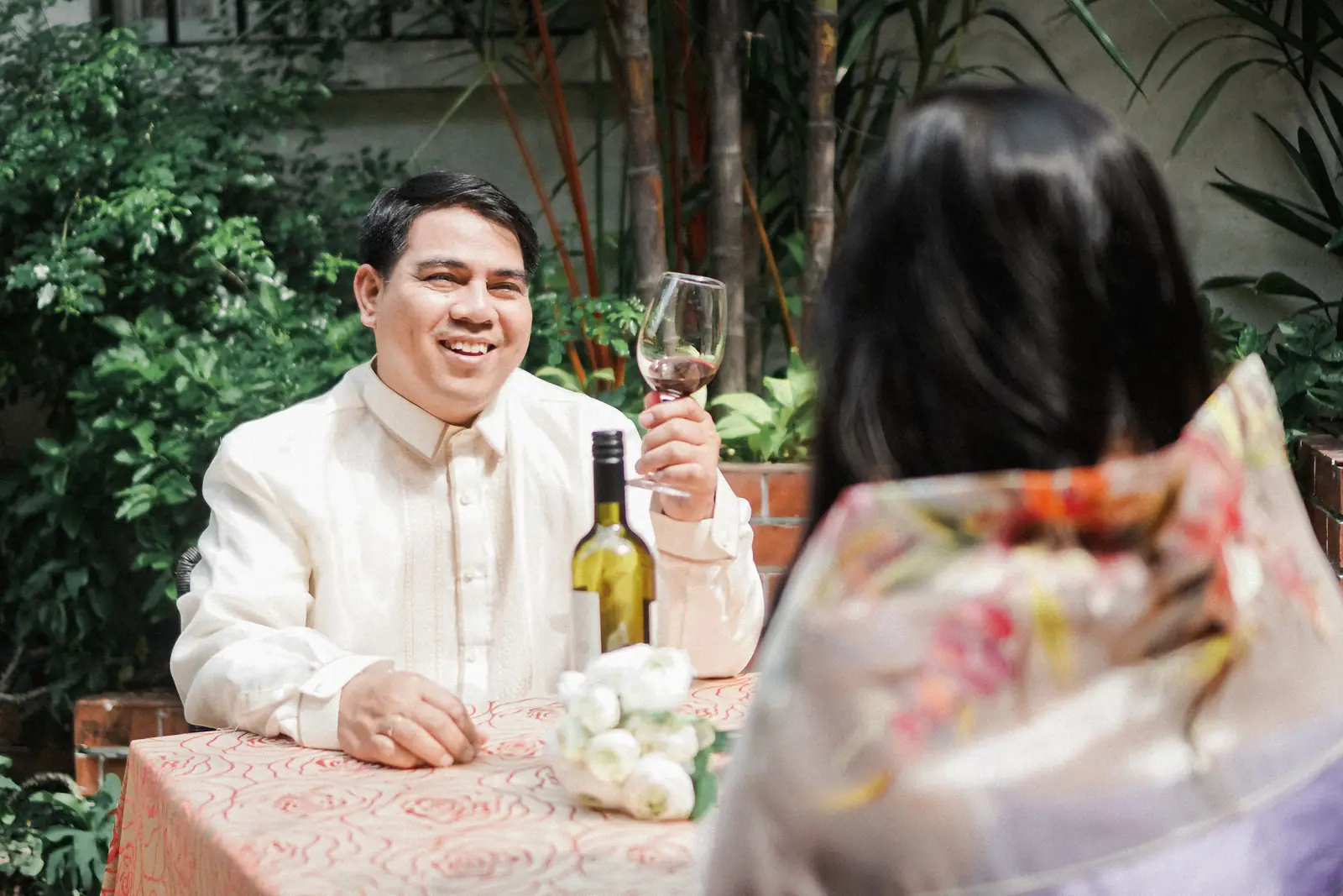 A smiling couple, he in a barong, at an outdoor garden table.