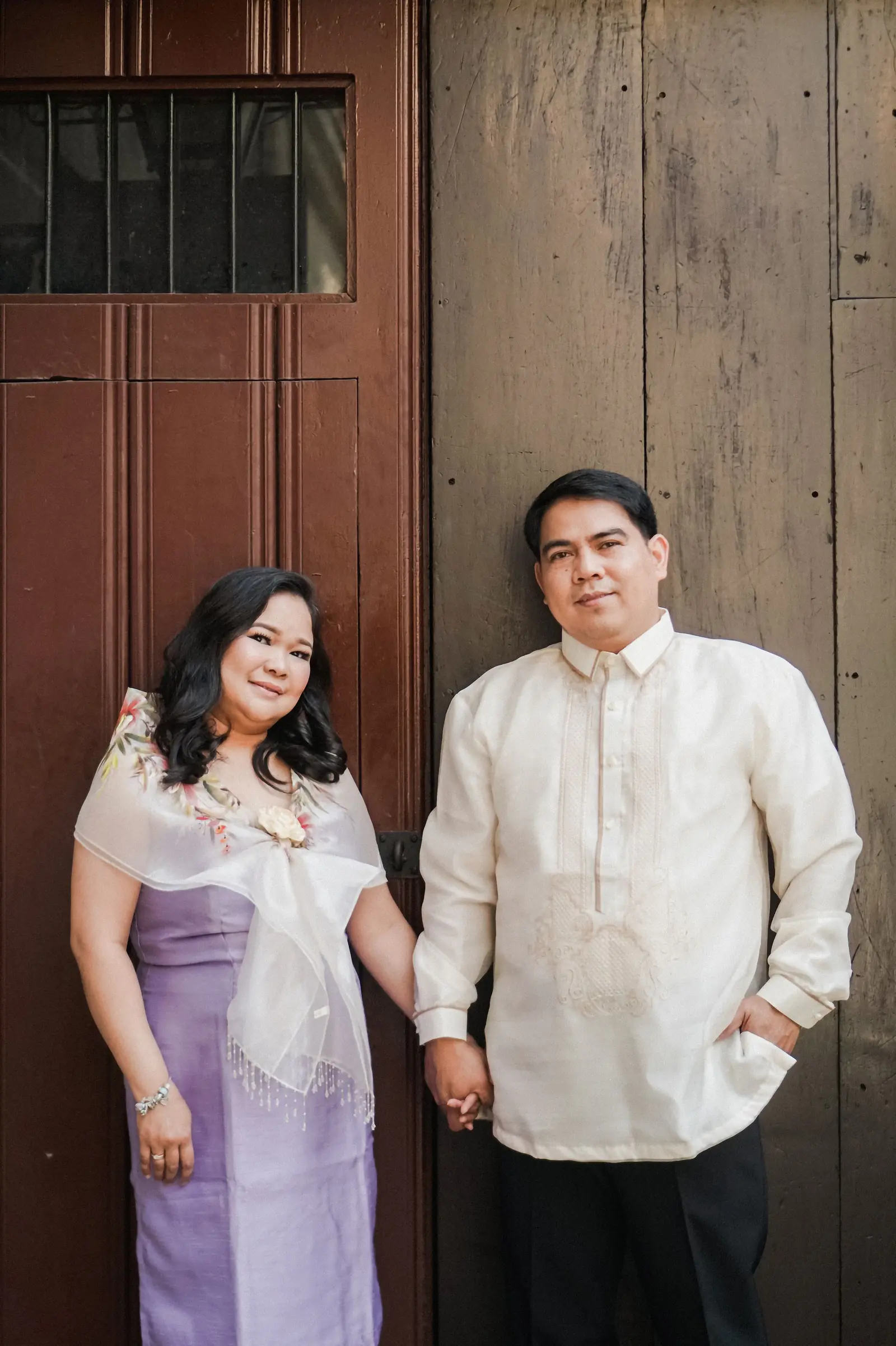 A couple in traditional attire holding hands against a wooden wall.