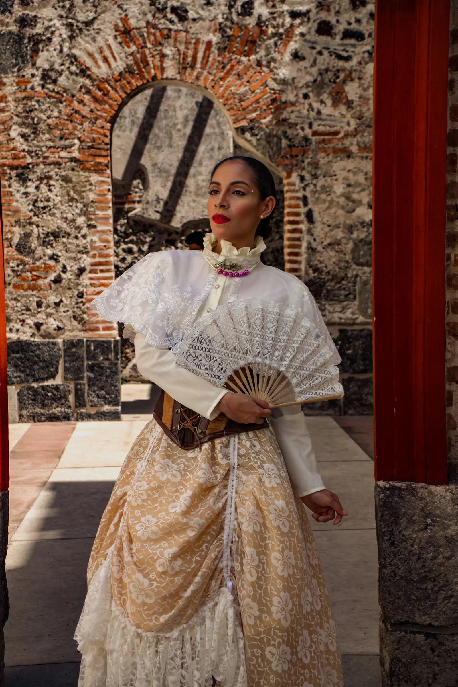 Woman in a traditional Filipiniana dress holding a fan in a historic setting.
