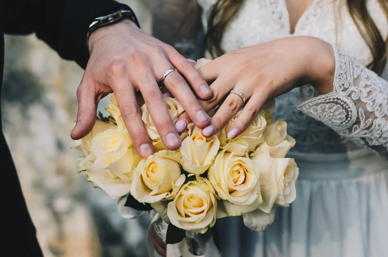 A couple's hands with wedding rings above a bouquet of yellow roses.